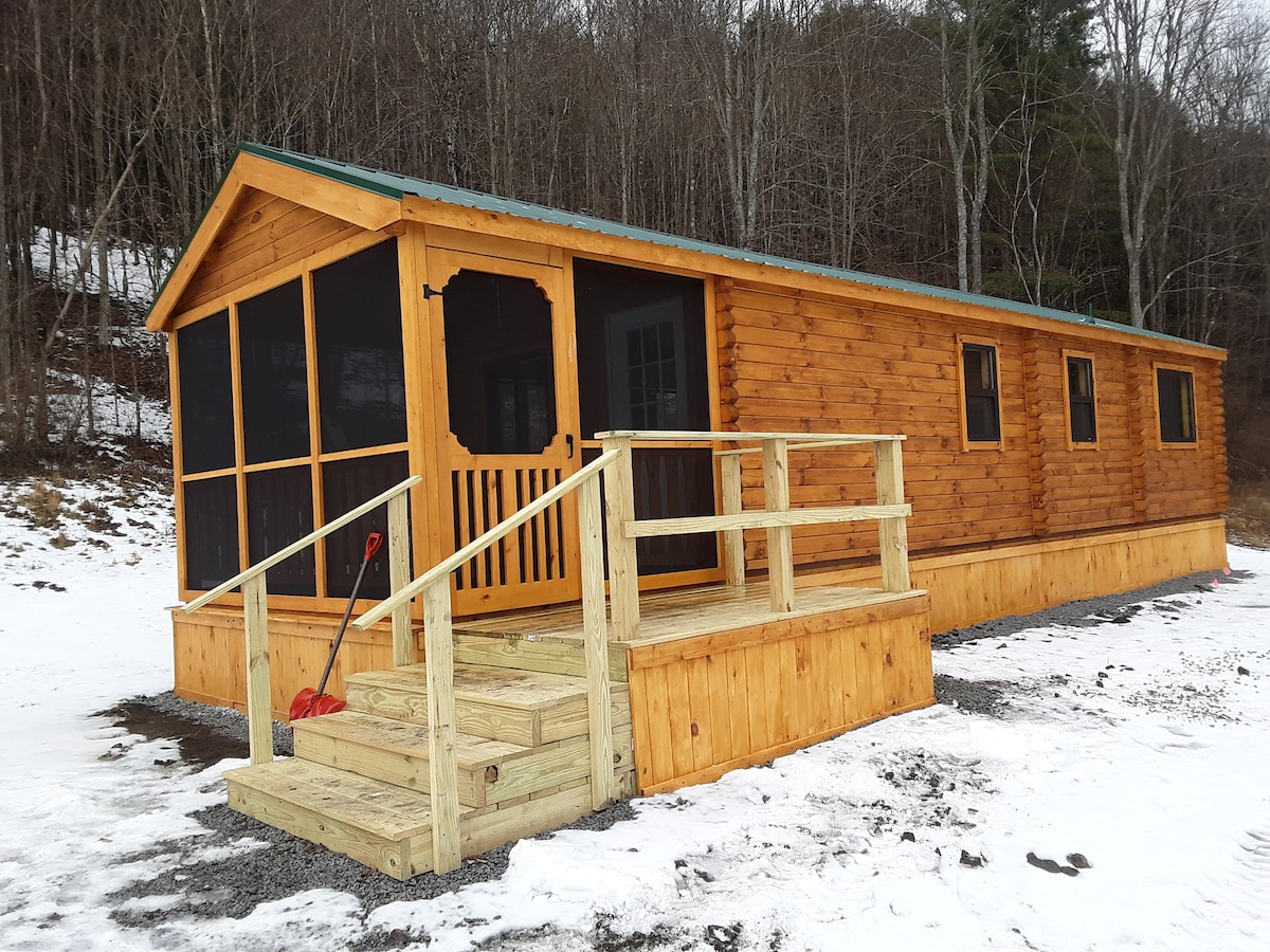 The exterior of a wooden cabin is showcased, featuring a screened-in porch with entrance steps. The cabin's warm wood tones contrast with the surrounding snowy landscape and trees. Large windows allow for natural light while the porch offers a space to enjoy the outdoors.