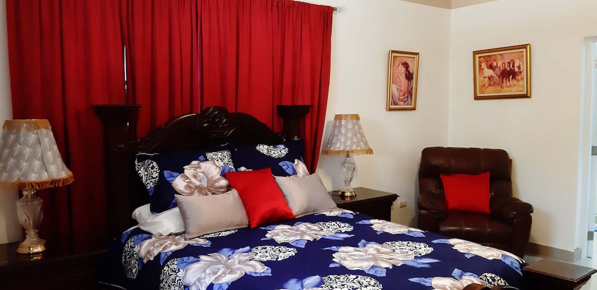 A furnished bedroom features a large bed dressed in floral-patterned linens, with red accent pillows. Two lamps with decorative shades stand on either side of the bed. A cozy armchair is positioned in the corner, complemented by a piece of wall art depicting horses.