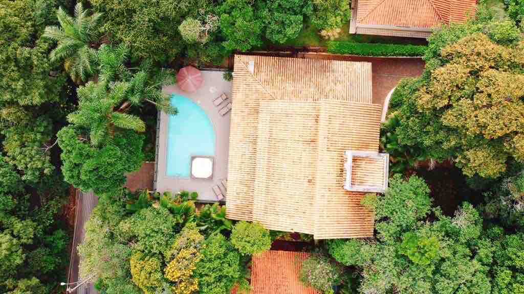An aerial view of a spacious home surrounded by lush greenery showcases a tranquil swimming pool with a sunbathing area. The residence features a textured roof that complements the natural landscape, creating a harmonious connection between building and environment.