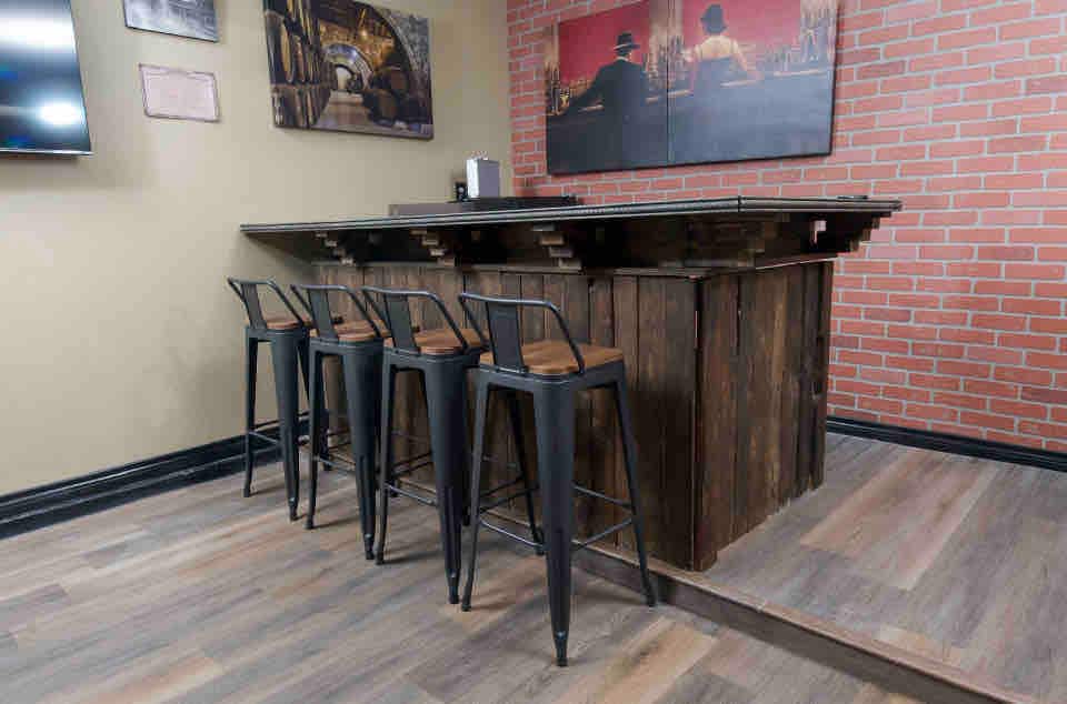 A rustic bar area is featured within the game room, highlighted by a wooden bar top and four metal stools with wooden seats. The wall is decorated with framed artwork depicting a speakeasy theme, complementing the inviting atmosphere of the space.