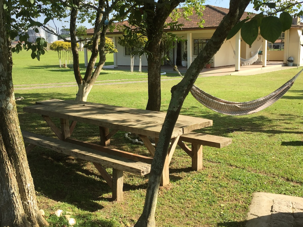 A wooden picnic table with attached benches is situated beneath trees, creating a shaded area on green grass. A hammock is visible nearby, inviting relaxation in the outdoor setting. The background features a stone path leading to a light-colored house.