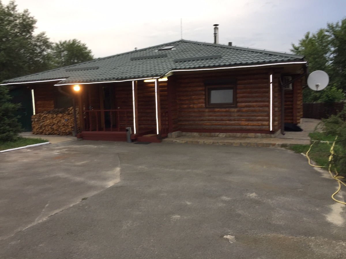 The wooden structure features a sloped roof and decorative lighting outlining the entrance. Stacks of firewood are visible nearby, and a satellite dish is mounted on the side. A spacious, paved area is present in front, suitable for parking and outdoor activities.