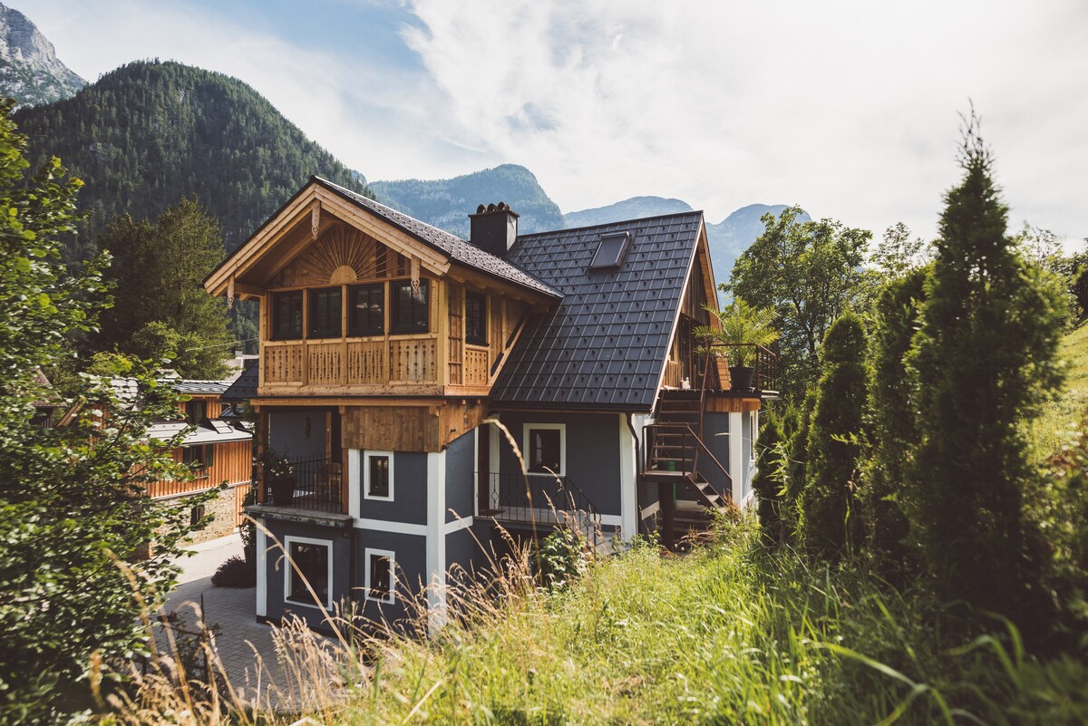 The exterior of a charming multi-level house is framed by lush greenery and mountains in the background. The building features a wooden balcony with decorative elements, a sloped roof, and a blend of natural materials, inviting guests to enjoy the serene surroundings.