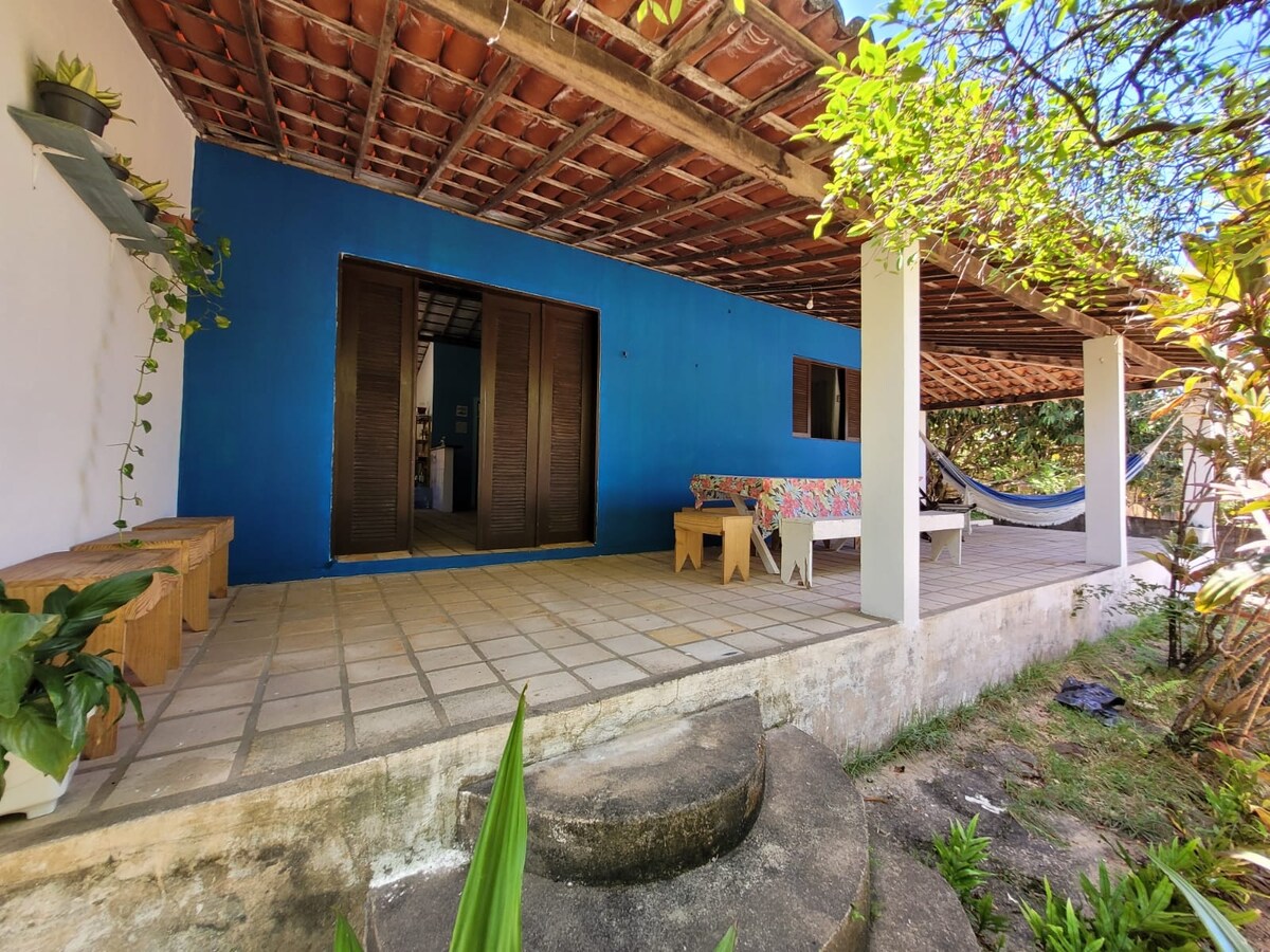 A spacious outdoor patio features tiled flooring under a wooden roof. Colorful seating arrangements are visible alongside a hammock. Lush green plants are placed throughout the area, providing a serene backdrop within a tranquil, natural setting.