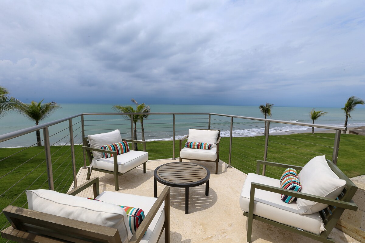 A private balcony features comfortable seating arrangements with four chairs and a round table, offering ocean views. Lush greenery surrounds the area, while the cloudy sky adds a serene ambiance. The sound of waves can be suggested by the distant shoreline.