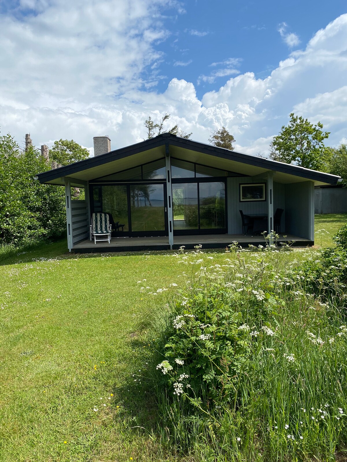 A charming summer house is situated on a grassy area, surrounded by greenery and wildflowers. Large windows provide views of the outdoors, while a covered porch features a rocking chair. Soft clouds drift across a blue sky, creating a serene atmosphere.