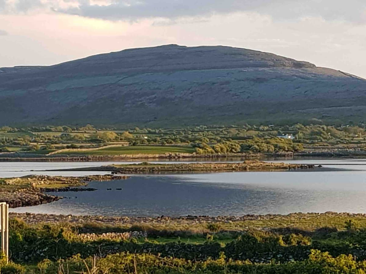 A sweeping view captures a distant mountain, its gentle slopes contrasting with the calm waters below. Lush greenery and a low stone wall frame the scene, reflecting the natural beauty of the surrounding landscape under a soft evening light.