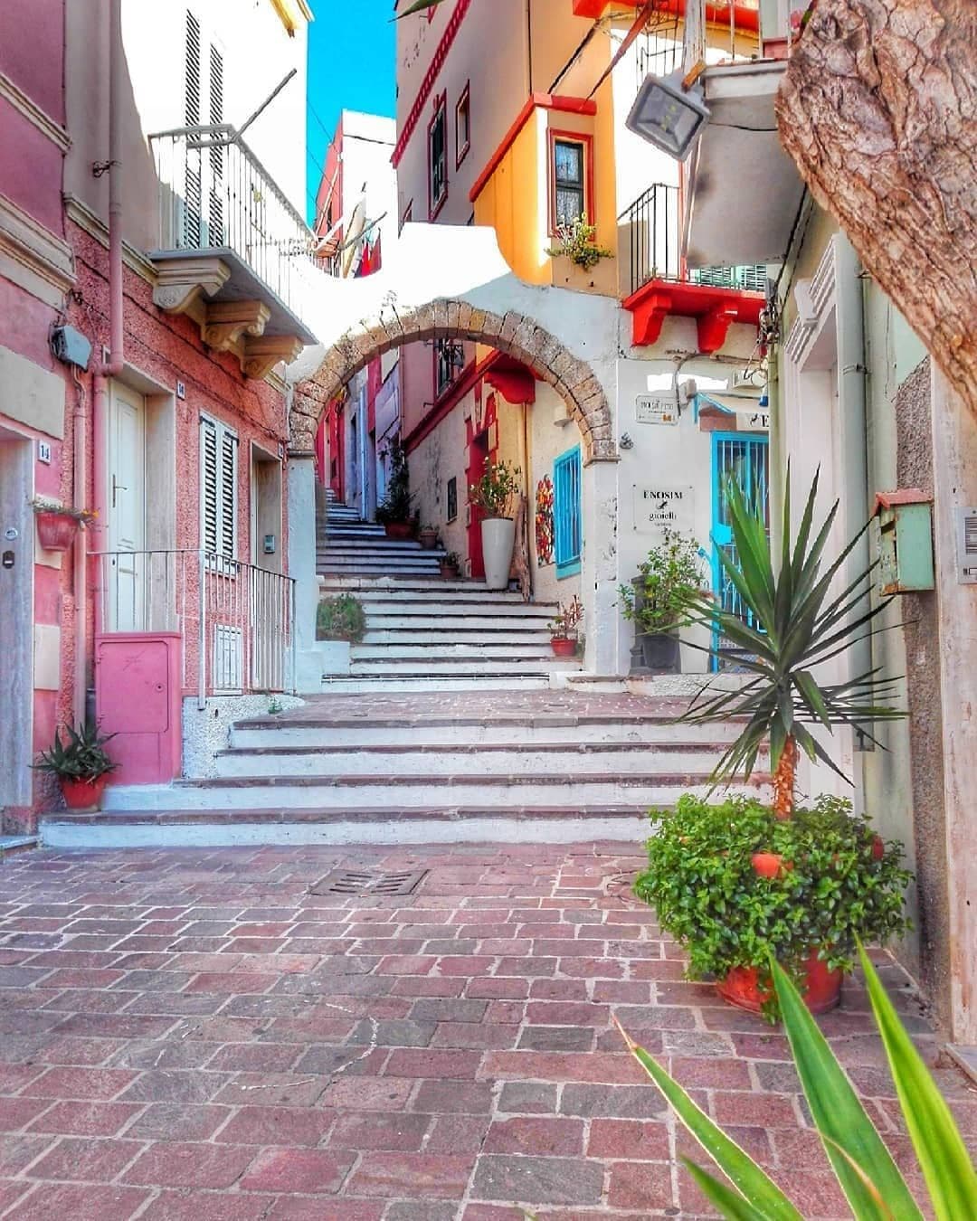 A charming, narrow street is shown, featuring a stone archway leading up a flight of steps. Colorful buildings line both sides, adorned with potted plants. The brick pathway invites exploration, while vibrant hues reflect the local architecture and ambience.