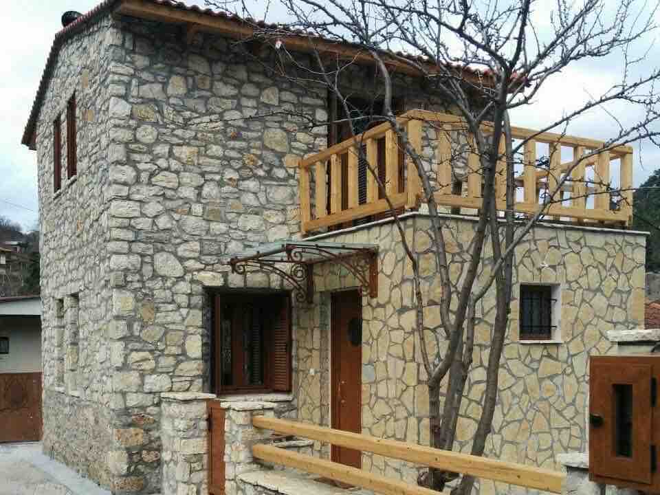 The exterior showcases a traditional stone house featuring a wooden balcony and railing. Large windows with wooden shutters complement the rustic design. The entrance is framed by a small, covered porch, and a tree adds a natural element to the setting.