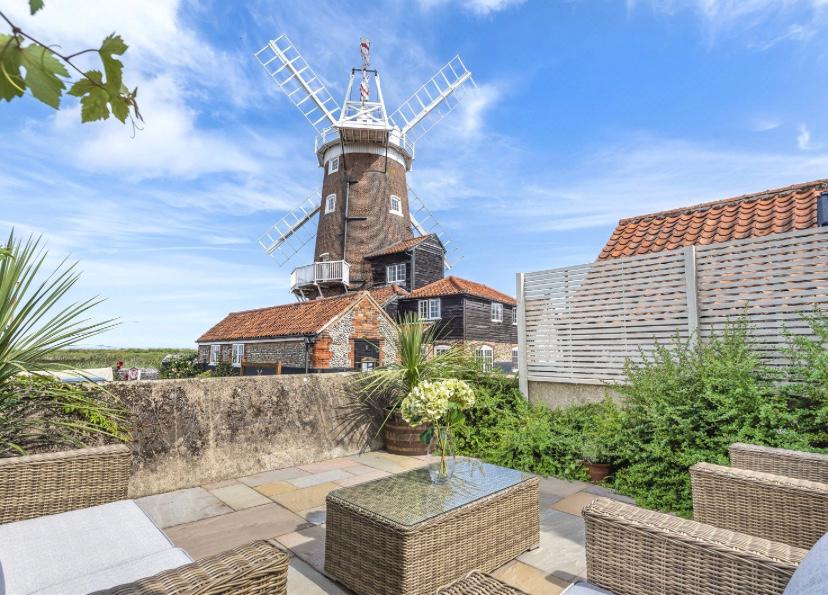 The outdoor seating area features a low table surrounded by wicker furniture, complemented by a backdrop of the historic Cley Windmill. Lush greenery and coastal vegetation frame the scene, with a clear blue sky adding to the serene atmosphere.