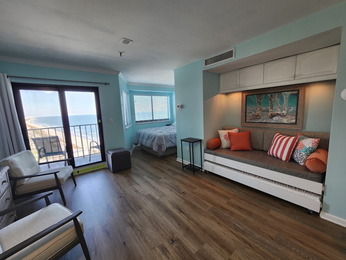 A bright studio space is featured with a queen bed in the background and a sleeper sofa in the foreground adorned with decorative pillows. Large windows provide natural light and a view of the ocean. A pair of chairs sits in the corner, adding to the calming atmosphere.