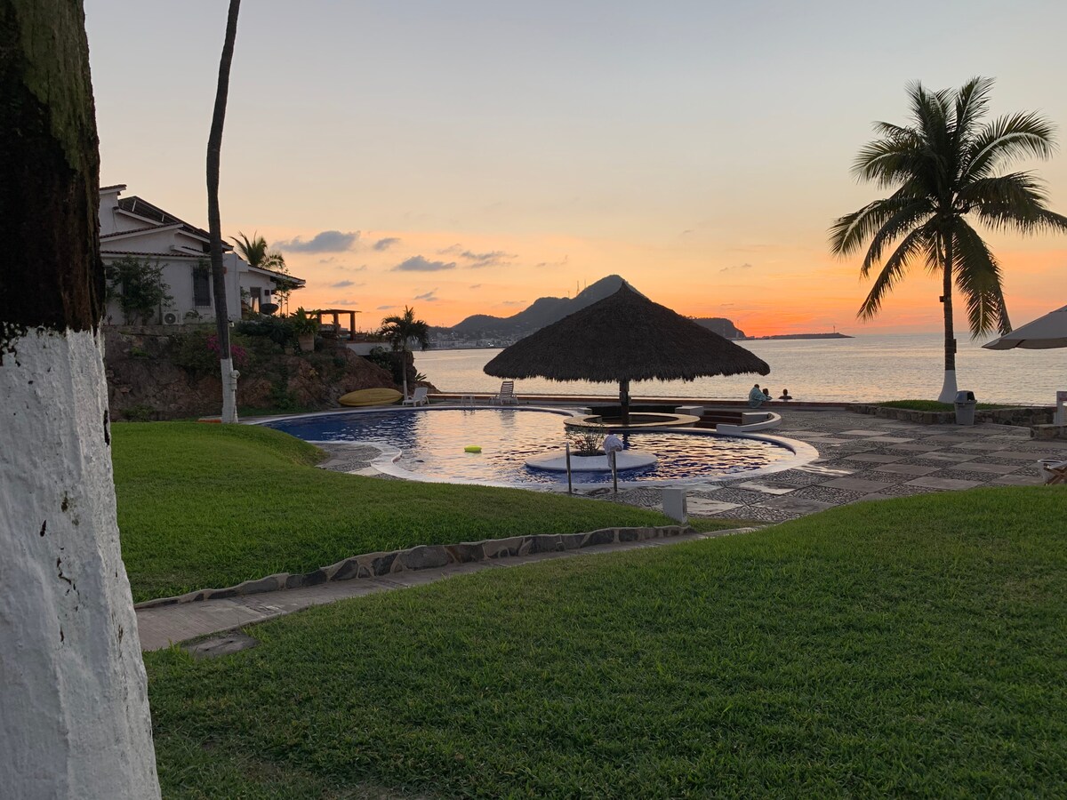 A tranquil pool area is set against a coastal backdrop, featuring a thatched palapa for shade. Lush green grass surrounds the pool, with palm trees framing the scene. The sunset casts warm hues over the water, enhancing the peaceful atmosphere.