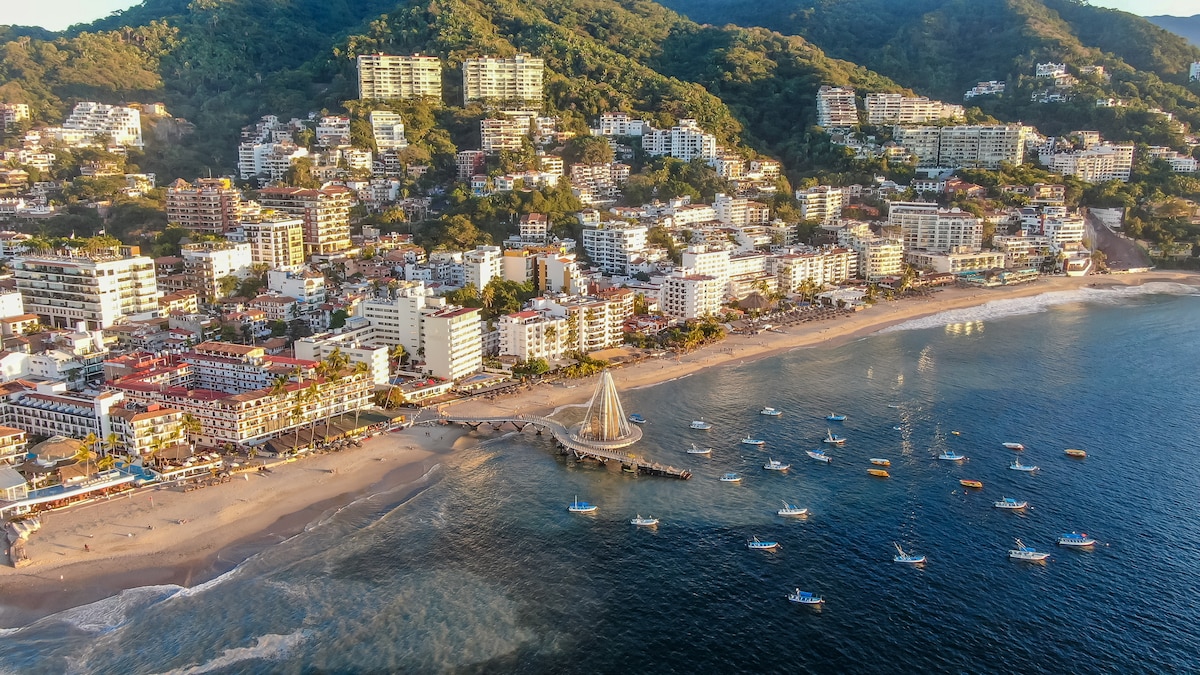 An aerial view captures a bustling coastal town, characterized by a sandy beach curving along the shoreline. Colorful buildings line the beachfront, with boats dotting the water. In the distance, green hills add a natural backdrop to the vibrant scene.