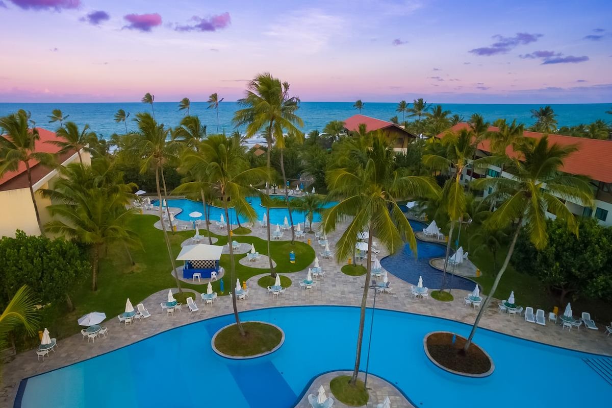 An expansive view of the resort's swimming pools is presented, featuring various shapes and shaded lounge areas. Lush palm trees surround the pools, contributing to a refreshing ambiance. The horizon showcases the ocean, with a twilight sky transitioning from blue to soft pink.