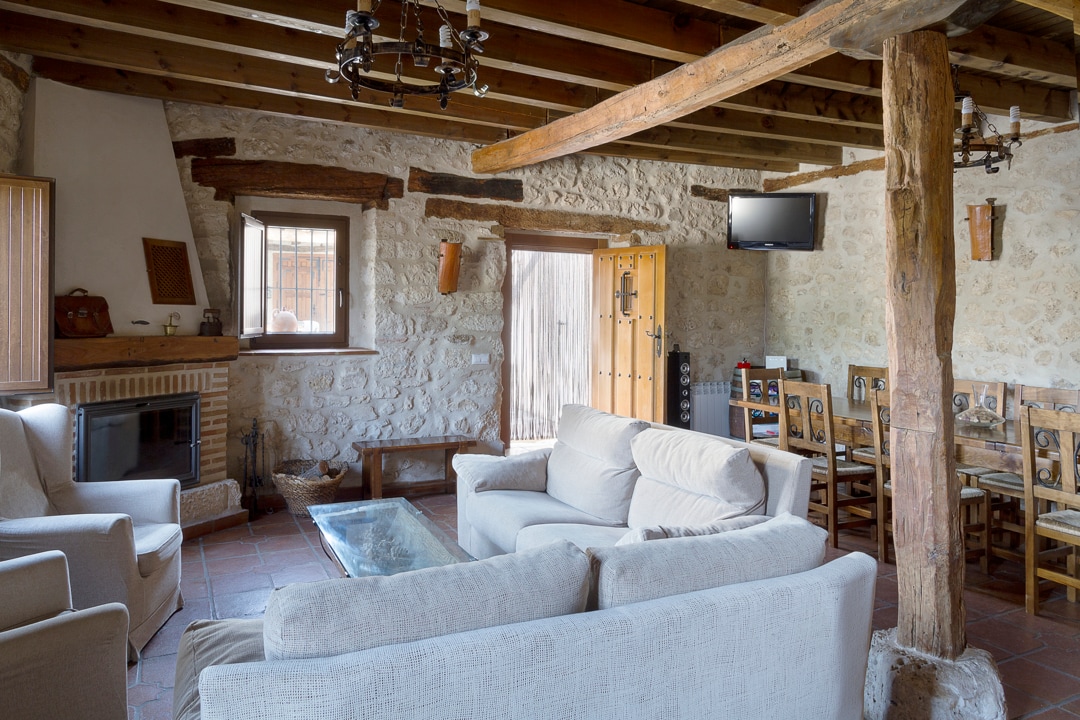 A spacious living area is highlighted by exposed wooden beams and a stone wall. Comfortable seating is arranged around a glass coffee table, with a wood-burning fireplace nearby. A dining area with wooden chairs is visible in the background, complementing the rustic ambiance.