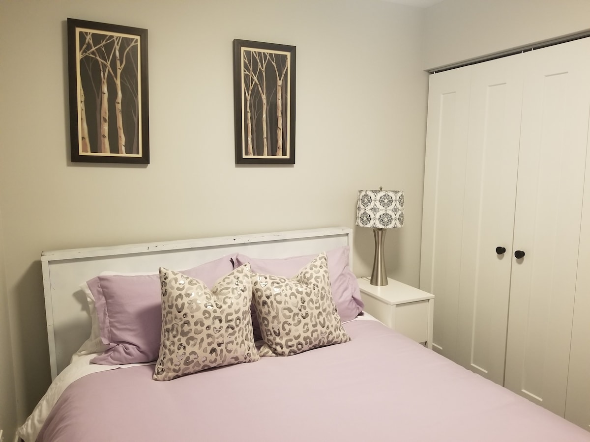 A calming bedroom features a bed adorned with purple linens and two decorative pillows with a leopard print. Framed artwork of trees hangs above the bed, while a stylish lamp sits on a nightstand beside it. White sliding doors provide storage space.