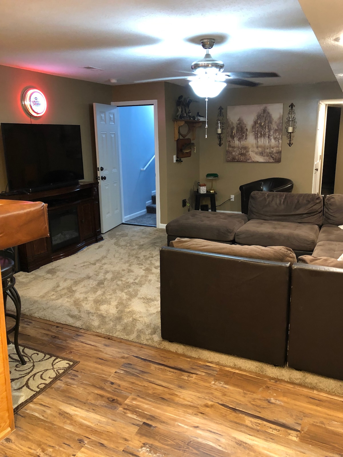 A cozy living area is presented with a large sectional sofa facing a flat-screen television. A ceiling fan emits soft light, while a staircase is visible in the background. The warm tones of wood and carpet create a comfortable and inviting space.
