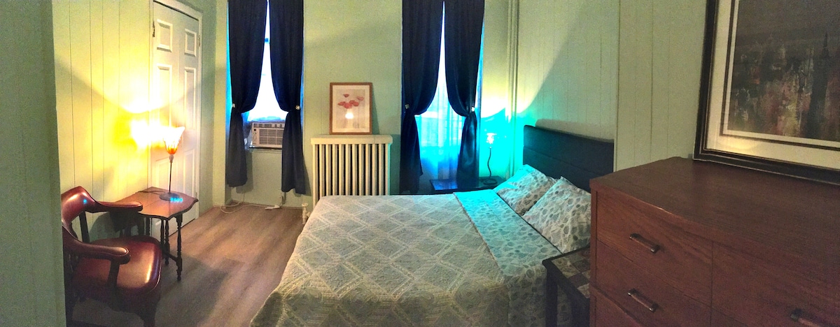 The bedroom features a spacious bed with patterned linens, complemented by a wooden dresser and a small table with a chair. A soft glow emanates from a bedside lamp, while blue curtains frame the windows, allowing light to enter. An air conditioning unit is positioned near the window.