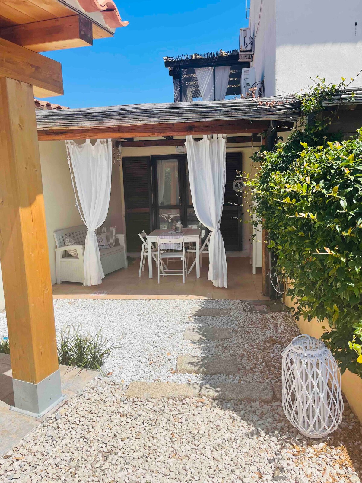 A charming outdoor area is visible, featuring a gravel pathway that leads to a shaded seating arrangement with a white table and chairs. Surrounding greenery and white drapes add a touch of privacy, while a decorative lantern contributes to the aesthetic.