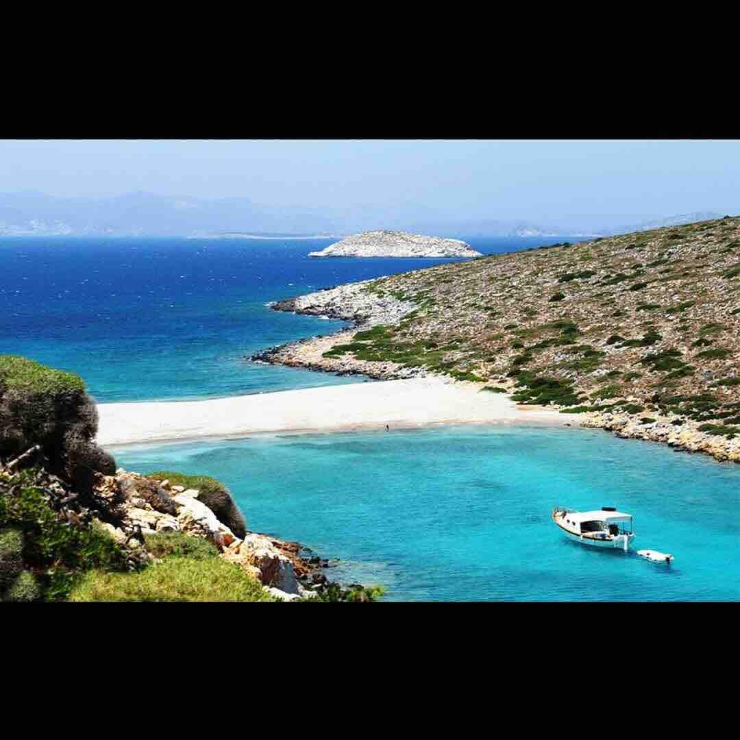 A serene coastline is depicted, featuring turquoise waters gently lapping against a sandy shore. Rugged hills with sparse vegetation rise on either side, leading to distant islands on the horizon under a clear blue sky.