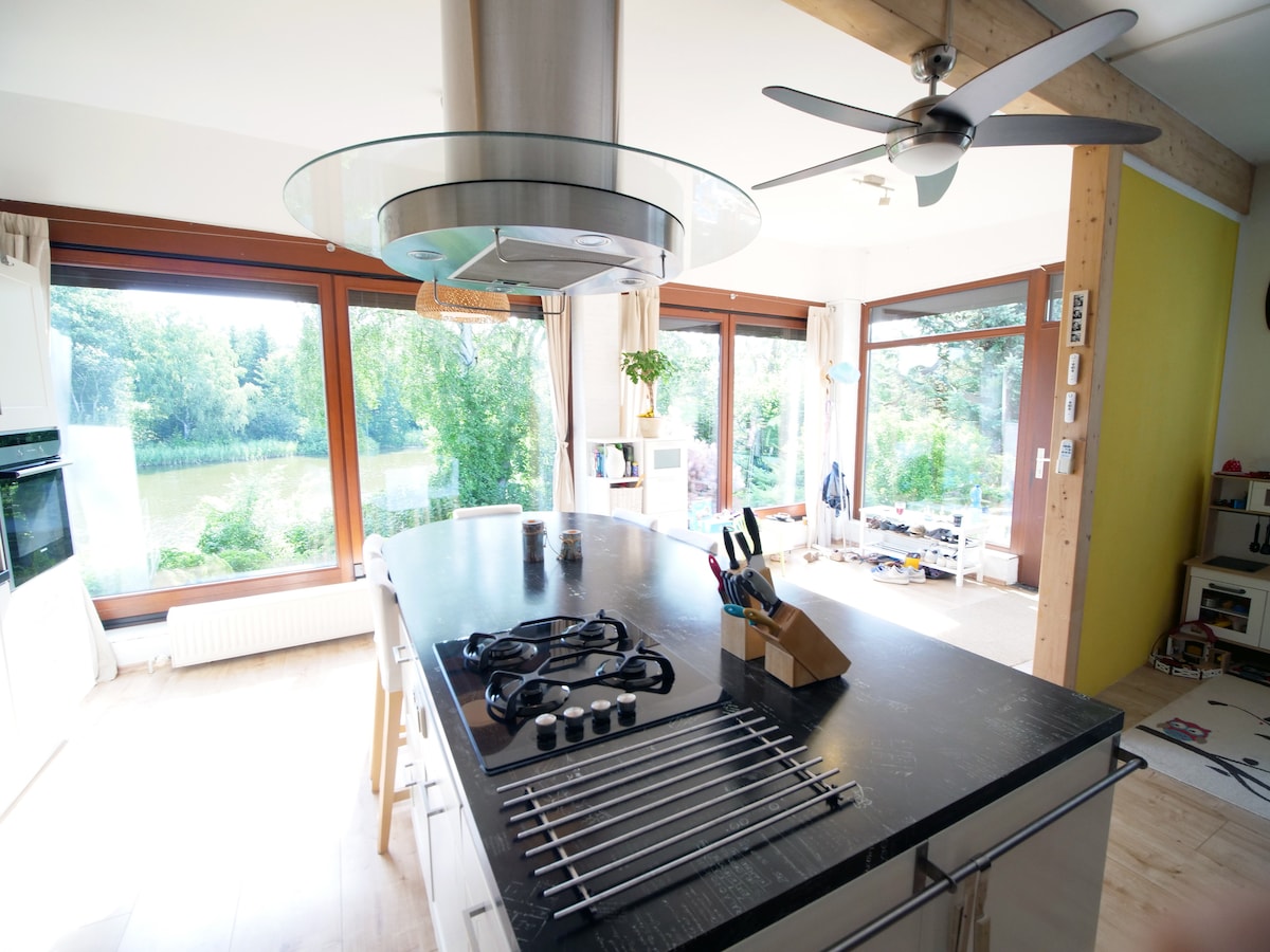 An open kitchen space features a large central island with a dark countertop and modern appliances. Large windows provide views of the surrounding greenery, while a ceiling fan contributes to the airy atmosphere. Kitchen utensils are neatly organized, enhancing functionality.