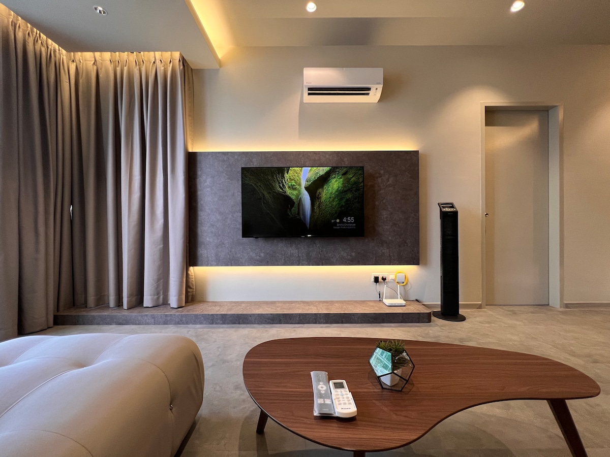 The living room features a modern design with a wall-mounted television displaying a nature scene. A sleek coffee table occupies the center, complemented by decorative items. Soft curtains frame a large window, while ambient lighting highlights the space's contemporary decor.