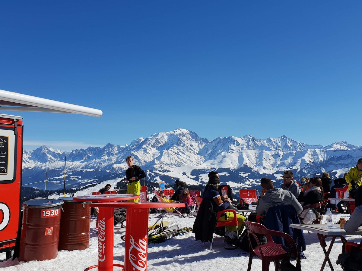 An outdoor seating area is visible, furnished with red chairs and tables atop snow-covered ground. A food truck stands nearby, with a scenic backdrop of snow-capped mountains under a clear blue sky. People are gathered, enjoying the vibrant alpine atmosphere.