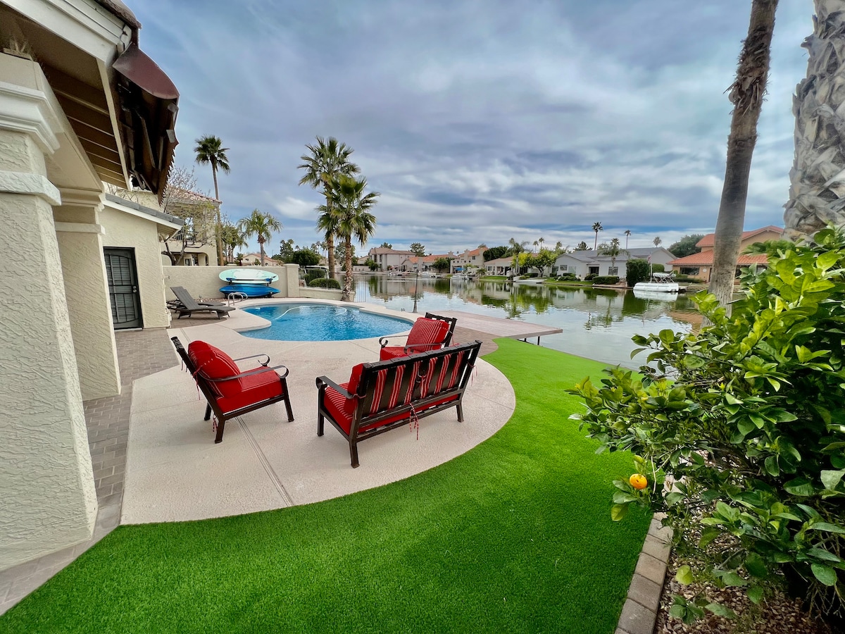 A serene outdoor area features a refreshing pool surrounded by lush, green landscaping. Two red lounge chairs are positioned near the water, offering a relaxing spot to enjoy the view of the lake and palm trees lining the shore.