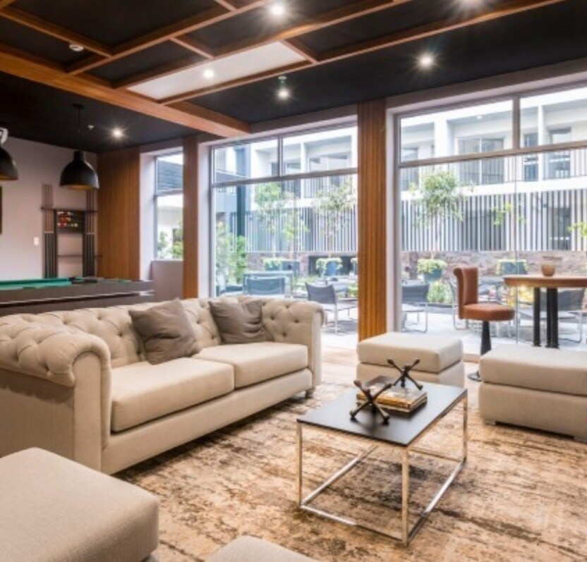 The communal lounge area features a stylish sectional sofa arranged around a low coffee table. Large glass doors provide views of an outdoor patio with seating. A billiards table and modern bar height tables are visible in the background, enhancing the inviting atmosphere.