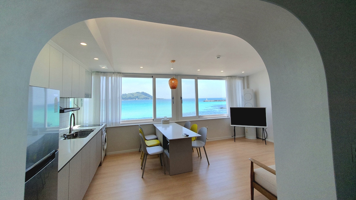 A spacious kitchen and dining area features large windows overlooking the turquoise sea and nearby island. A modern dining table is surrounded by grey chairs, while a cozy seating area includes a light wood frame chair. Natural light illuminates the bright, open space.