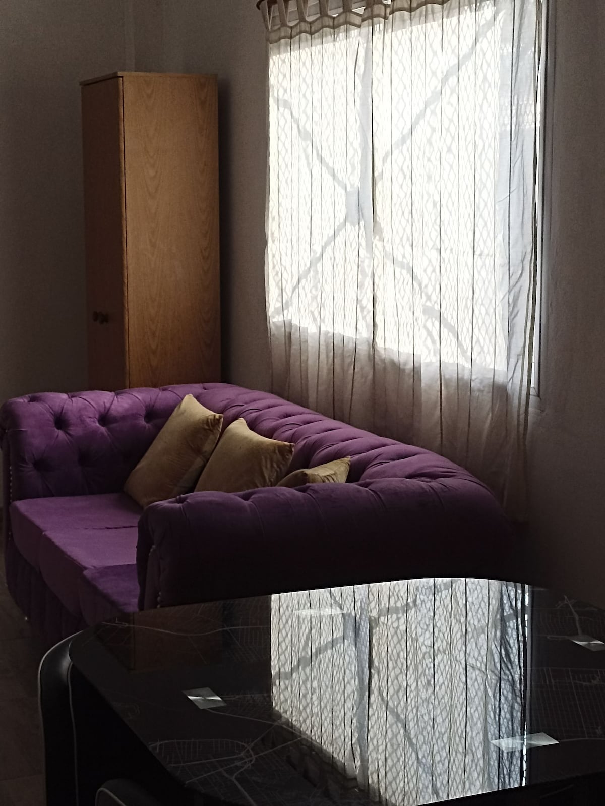 A plush purple sofa is adorned with decorative cushions, positioned beside a window dressed with sheer curtains. Light filters in, casting soft reflections on the smooth, dark surface of a nearby table. A wooden cabinet stands against the wall.