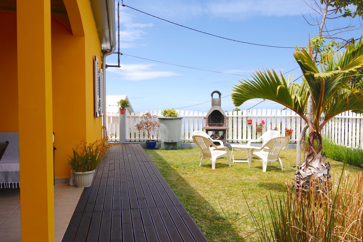 The outdoor terrace features a wooden deck leading to a grassy yard. Two lounge chairs are positioned near a barbecue area. Bright yellow exterior walls complement the setting, with potted plants adding greenery along the fenced perimeter. The ocean view is visible in the background.