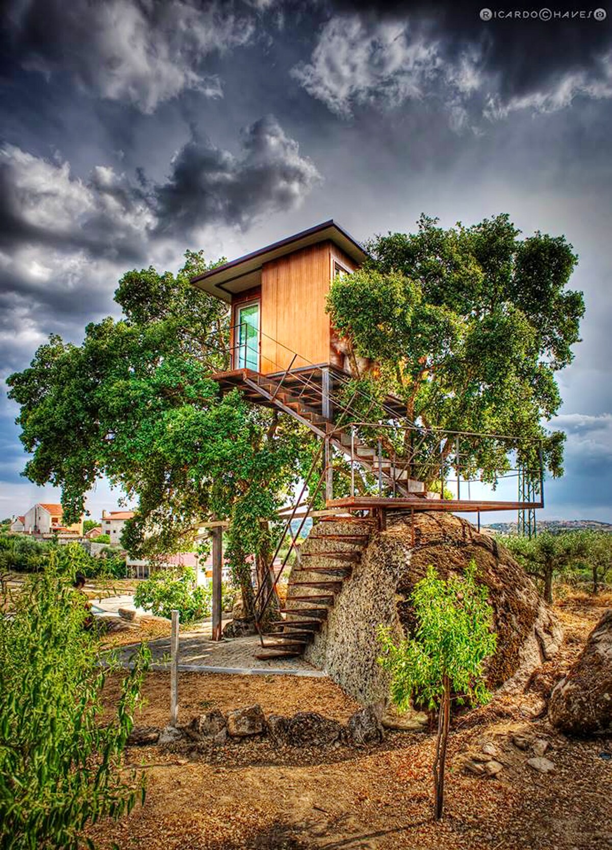 A modern treehouse is set among lush greenery, elevated on a rocky base. Sturdy stairs lead to the entrance, which features large windows allowing natural light to fill the interior. The surrounding landscape provides a blend of trees and open space.