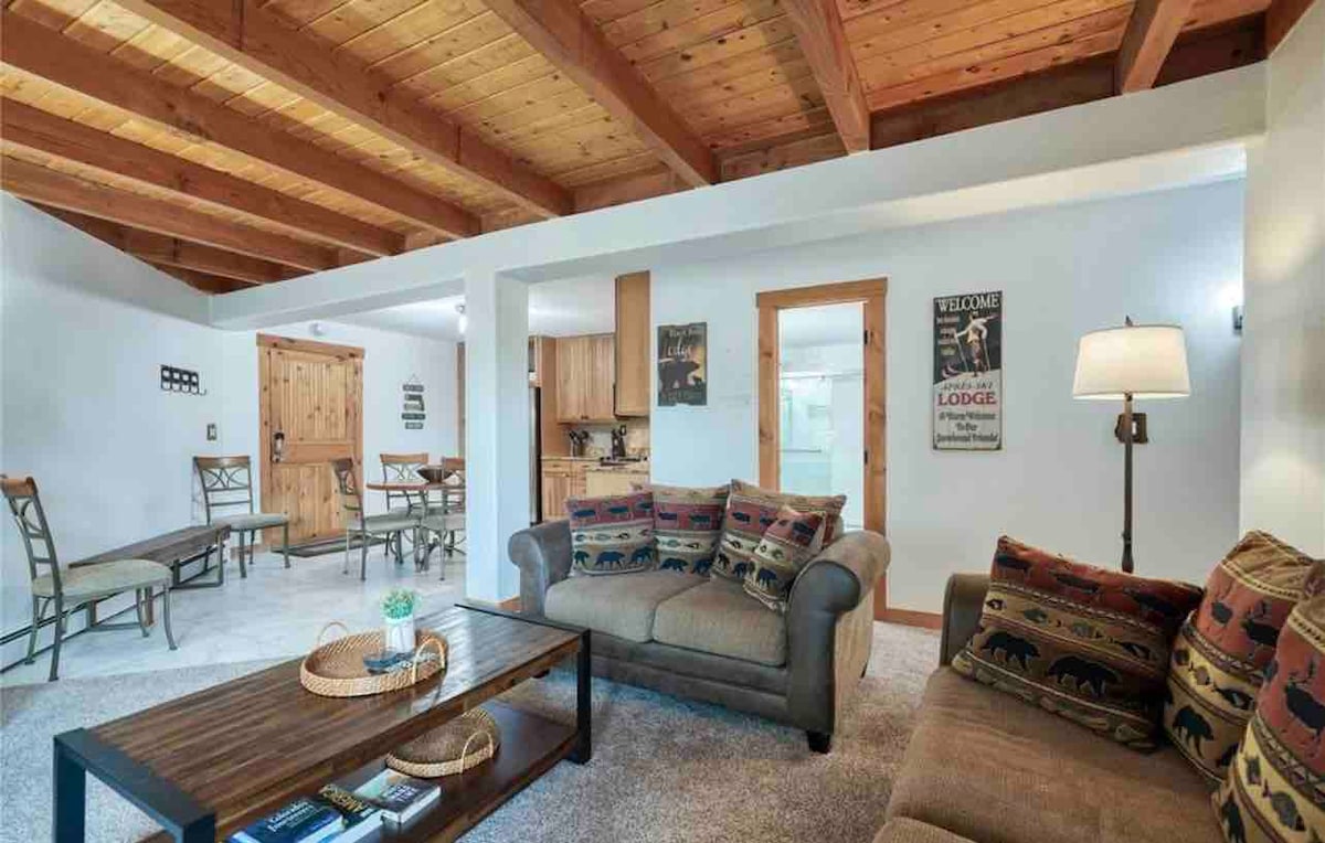 A well-lit living area features a cozy sofa adorned with patterned cushions. Rustic wooden beams span the ceiling. A coffee table sits centrally, displaying a decorative tray. A dining area with a table and chairs can be seen in the background, contributing to the open layout.