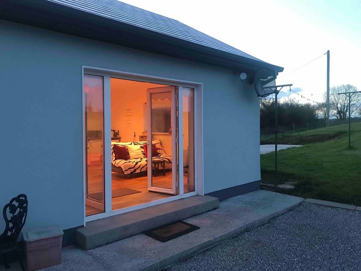 The exterior of the chalet is shown at dusk, featuring large glass doors that reveal a warmly lit interior. A welcoming entryway leads to a cozy living space, while a grassy area extends beyond. Outdoor seating is visible alongside the pathway.