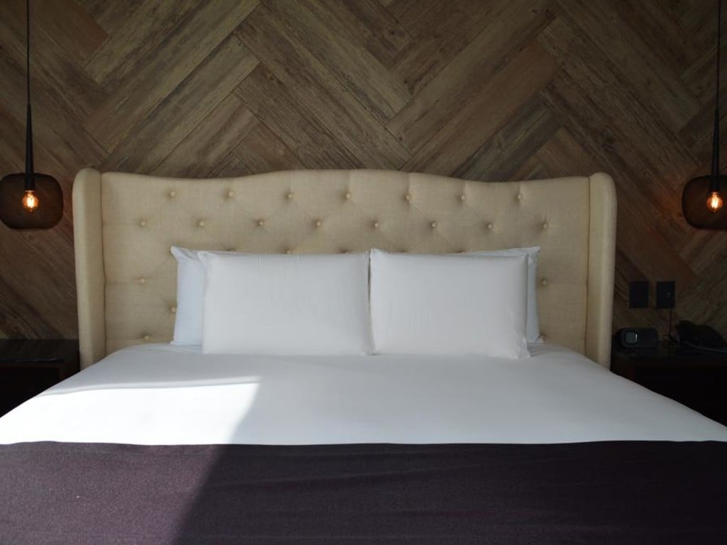The spacious bed features a tufted headboard and is adorned with crisp white pillows. Soft lighting is provided by hanging fixtures on either side. Natural wood walls create a warm and inviting ambiance.