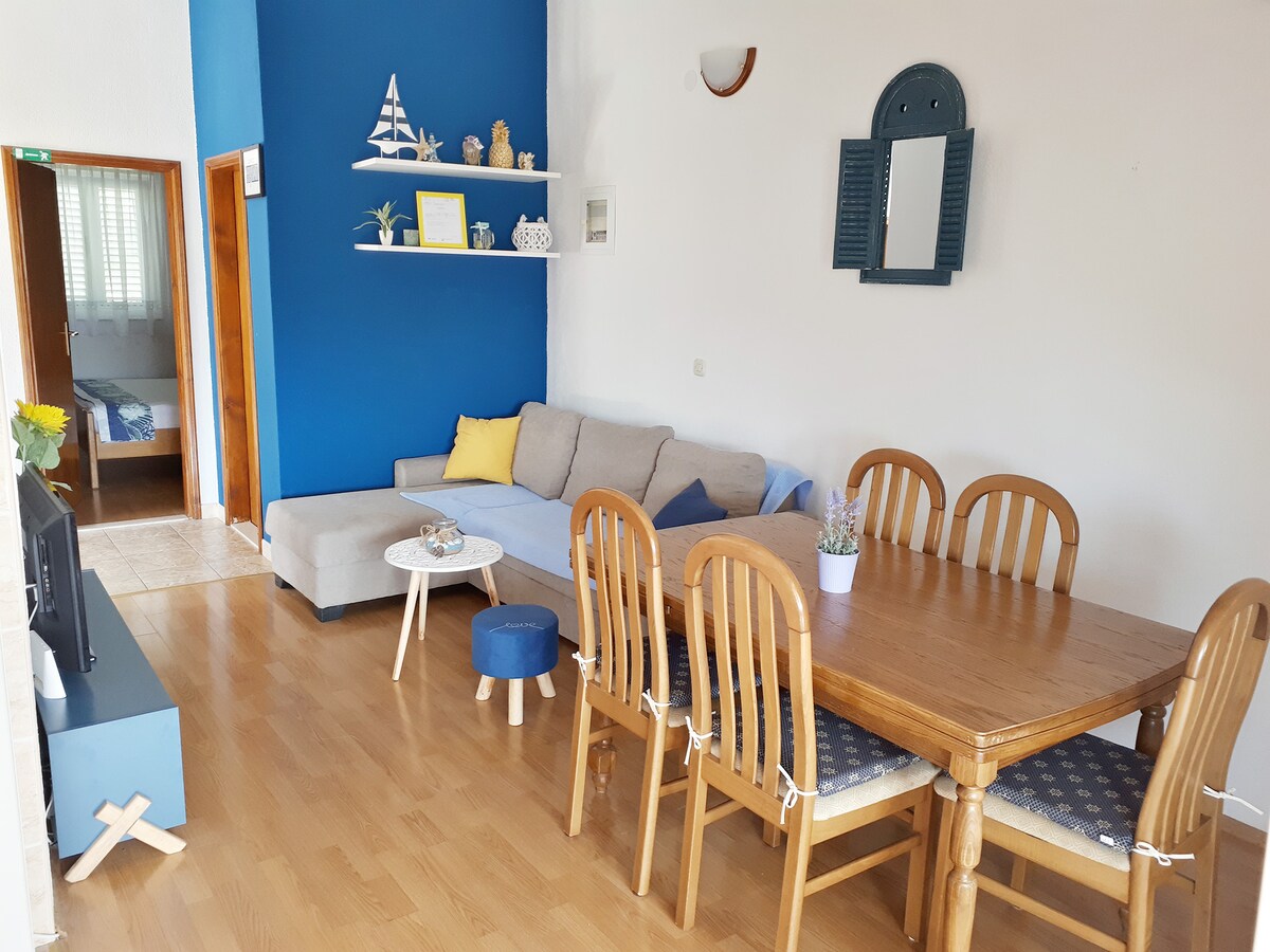 A spacious living area features a wooden dining table surrounded by chairs and a comfortable seating arrangement with soft cushions. A blue accent wall provides contrast, while a shelf displays decorative items. Light enters through a nearby window, enhancing the welcoming atmosphere.