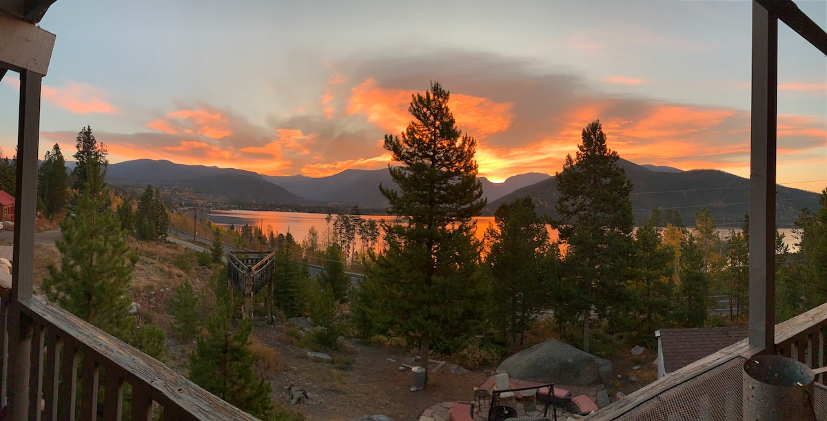 A panoramic view from the wrap-around porch showcases a vibrant sunset over Shadow Mountain Lake and surrounding mountains. The sky displays shades of orange and pink, while the serene water reflects the evening colors. Tall trees frame the scene, enhancing the natural beauty.