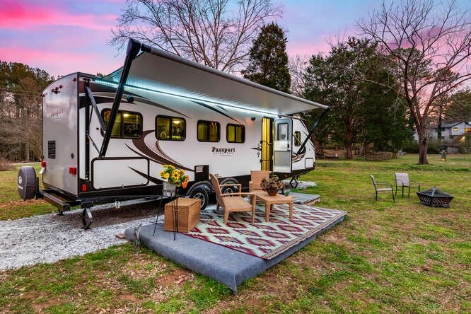 A luxury RV is shown parked on a grassy area, surrounded by trees. An outdoor rug is laid down, complemented by wicker furniture and a firepit. The awning provides shade over the seating area, while a vibrant floral arrangement adds a touch of color.
