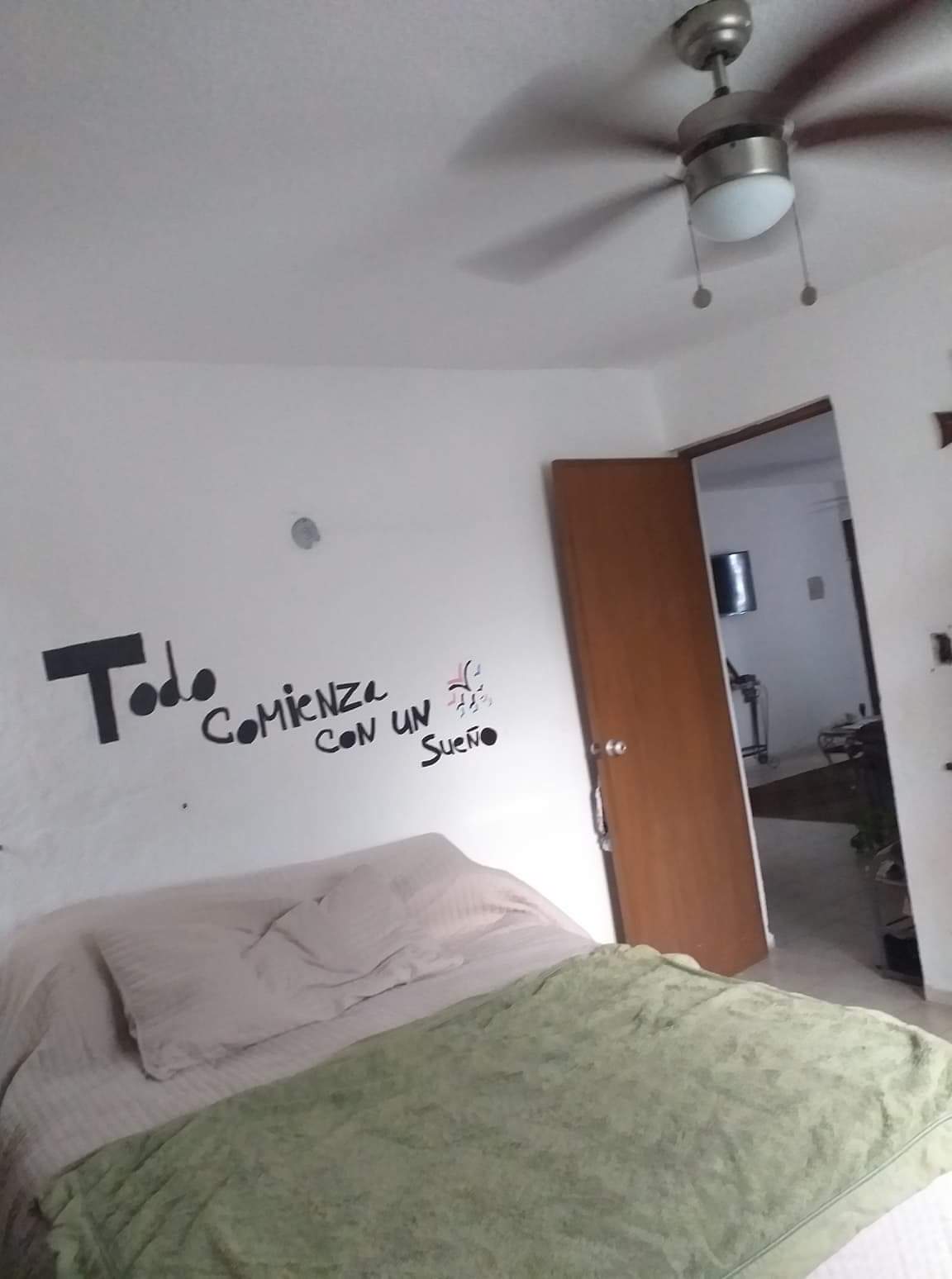 A simple sleeping area features a spacious bed with light bedding and a green blanket. A ceiling fan is mounted above, and the walls are painted in soft white. An open door leads to another room, creating an inviting openness.