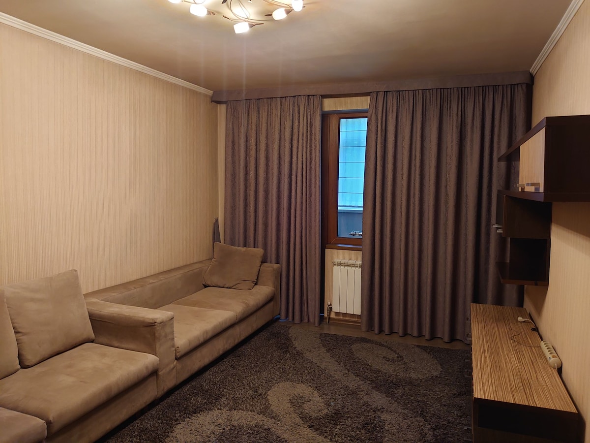 A well-lit living area features a plush sectional sofa and a flat-screen television mounted on a wooden unit. Tall curtains frame the window, which allows natural light to filter in. A soft, patterned rug covers the floor, creating a cozy atmosphere.