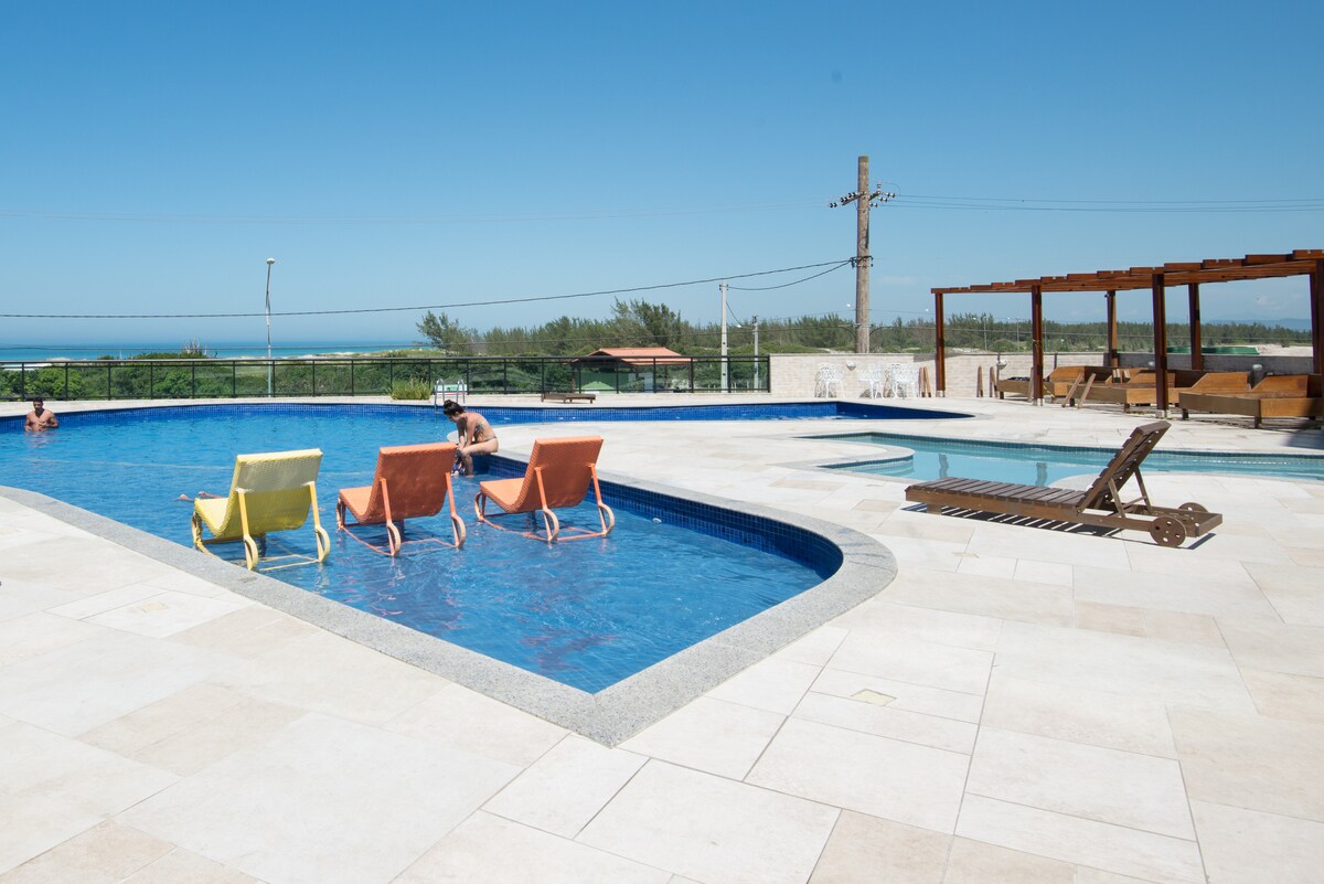 A spacious outdoor pool area features various loungers positioned around the water. The blue pool reflects sunlight under a clear sky, while a sauna and shaded seating are visible in the background, providing options for relaxation.