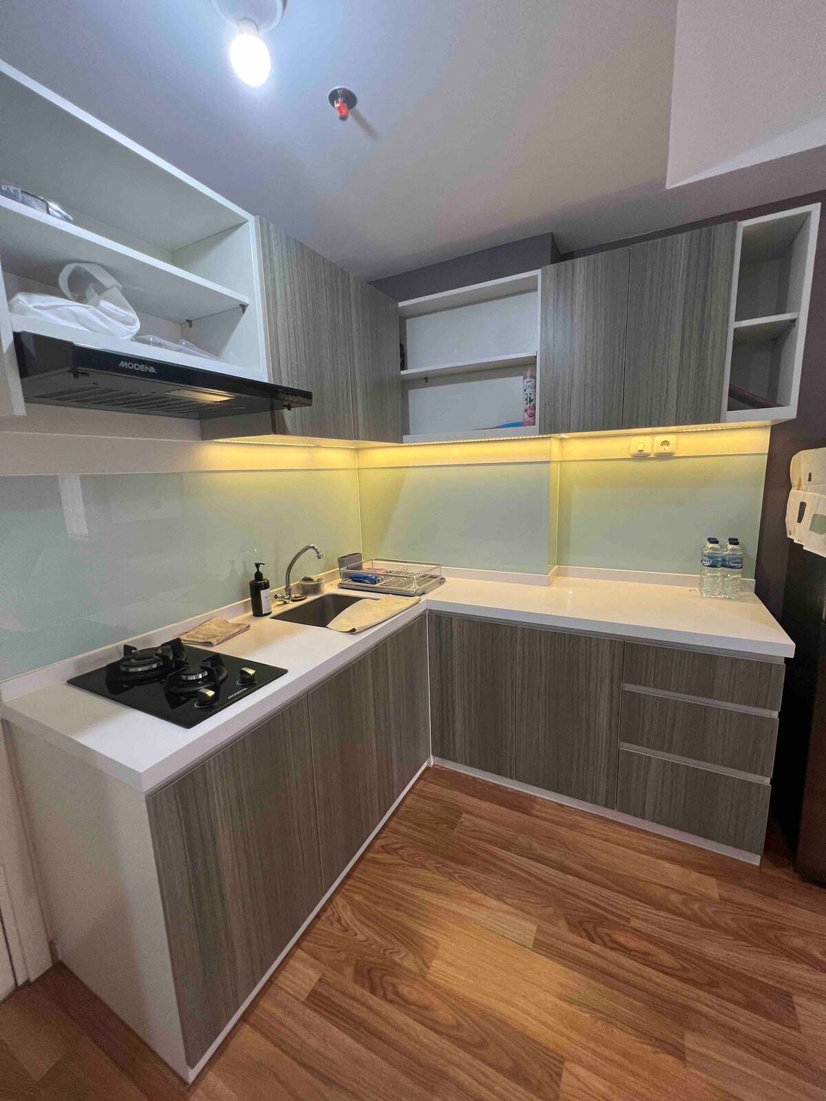 A modern kitchen features sleek cabinetry in neutral tones, complemented by a reflective backsplash. Stainless steel appliances include a hood and a stovetop. Natural light enhances the workspace, while warm lighting creates a functional area for meal preparation.