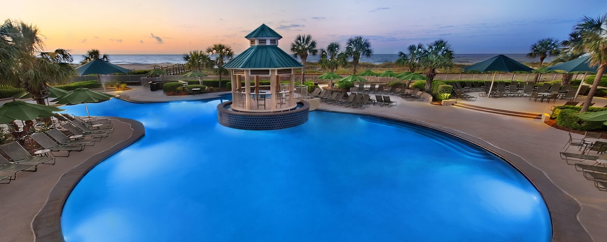 A large swimming pool is surrounded by palm trees and lounge chairs. A gazebo with a green roof sits at the center of the pool. The ocean and a sunset sky are visible in the background, creating a serene setting.