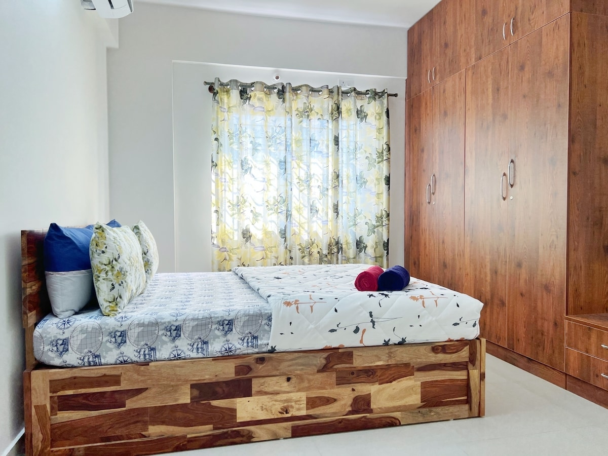 A serene bedroom features a spacious wooden bed with a floral-patterned mattress and two decorative pillows. Natural light flows through floral curtains, brightening the light-colored walls. A sturdy wooden wardrobe stands gracefully against the wall, adding ample storage space.