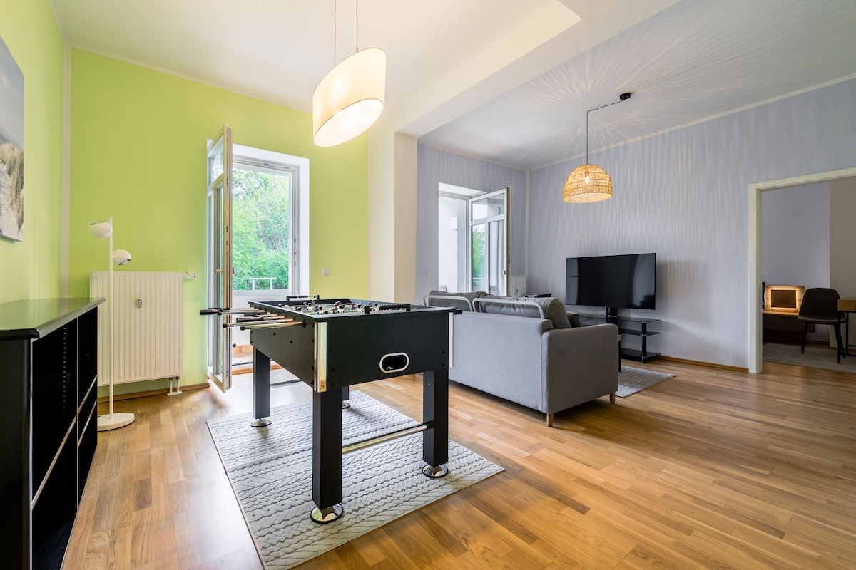 A spacious living area features a dark foosball table at the center, surrounded by light wood flooring. Two large windows provide natural light, highlighting a comfortable gray sofa and a flatscreen TV mounted on the wall. A warm glow is emitted from a modern light fixture overhead.