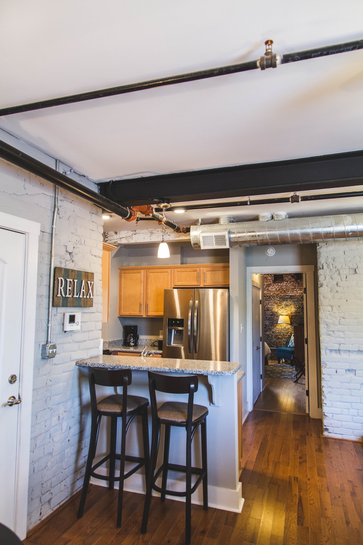 A well-equipped kitchen area features wooden cabinets and a granite breakfast bar with two stools. Industrial pipes are visible along the ceiling, complementing the exposed brick walls. A doorway leads to a cozy living space, while a sign reading 'RELAX' adds a welcoming touch.