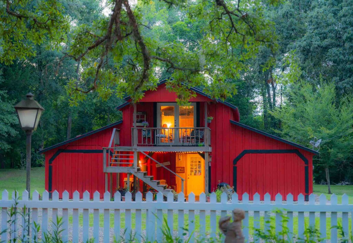 A charming red barn loft is embraced by lush greenery, with a white picket fence framing the view. The exterior features large windows that illuminate the interior. A staircase leads to the entrance, offering a welcoming perspective of the property nestled among the trees.
