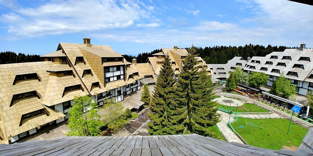 An aerial view of the apartment complex is presented, highlighting the distinct wooden architecture with sloped roofs. Surrounding green spaces and trees appear well-maintained, offering a sense of tranquility. The central courtyard features pathways and a grassy area, inviting relaxation and outdoor activities.