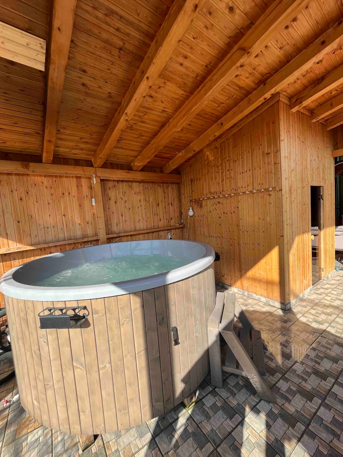 A wood-paneled area features a round hot tub surrounded by natural light, accentuated by the warm tones of the wooden ceiling. Steps lead up to the tub, providing easy access. The tiled floor enhances the inviting ambiance of the space.